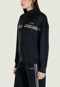 Felpa nera con zip e collo alto, con strisce di strass sul petto e due tasche laterali. Texture liscia e design minimalista.