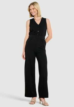 Femme souriante aux cheveux blonds portant une combinaison noire sans manches avec ceinture et sandales à talons hauts dorés, debout devant un fond uni.