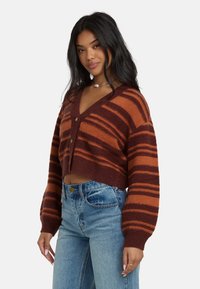 Braun-orange gestreifter, verkürzter Cardigan mit V-Ausschnitt, Knopfverschluss und weichem Material. Kombiniert mit hoch taillierten blauen Jeans.