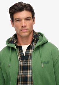Groene zip-up hoodie met trekkoord en logo, gedragen over een marineblauw- en groen geruit overhemd met knopen. Gladde textuur en een ontspannen pasvorm.