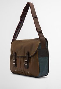 Καφέ καμβάς τσάντα messenger με δερμάτινη επένδυση, ρυθμιζόμενος πολυχρωμος ριγωτός ιμάντας και δύο ασημένιες πόρπες στο μπροστινό μέρος.