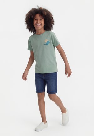 Copil zâmbitor cu păr creț, purtând un tricou verde cu mâneci scurte, pantaloni scurți din denim și pantofi albi slip-on, mergând înainte pe un fundal alb.