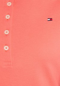 Corallinvärinen polo-paita, joka on valmistettu tekstuuri-kankaasta. Paidassa on kolme nappia edessä ja pieni brodeerattu logo punaisena, valkoisena ja sinisenä.