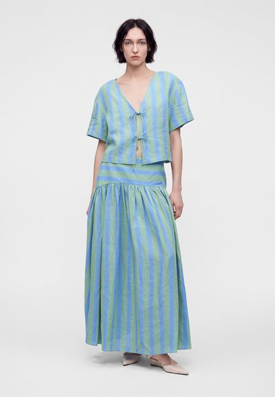 Femme portant un haut à manches courtes rayé vertical bleu et vert noué à l'avant, ainsi qu'une longue jupe fluide assortie avec des chaussures blanches pointues.