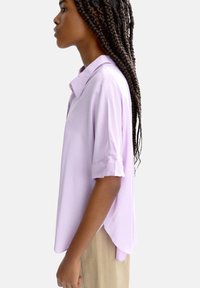 Jasnofioletowa bluzka z krótkim rękawem, z kołnierzykiem, gładka w fakturze, z nieco dłuższym tyłem, w połączeniu z beżowymi spodniami.