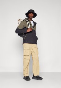 Schwarze und olivgrüne Zip-Jacke, beiger Cargo-Hose, schwarze Sneaker und ein schwarzer Bucket Hat. Der Rucksack hat hellfarbige Träger.