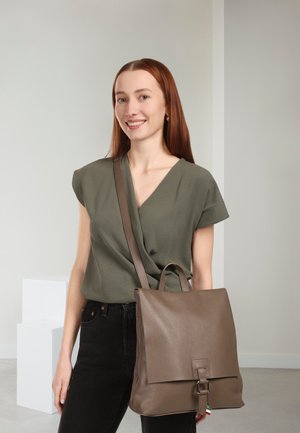 Sac à dos en cuir marron à finition texturée, forme rectangulaire et bretelles réglables. Porté sur un haut vert, avec une silhouette fine.