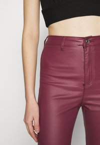 Pantalon en cuir synthétique bordeaux à la coupe ajustée, doté d'une fermeture éclair et d'un bouton à l'avant, avec un léger éclat. Détails de couture minimaux visibles.
