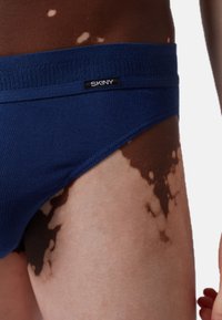 Briefs da uomo blu navy con una texture a coste e un elastico con un'etichetta nera. La pelle esposta mostra una pigmentazione marrone e a fantasia chiara.