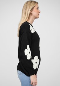 Pull en tricot noir avec de grandes motifs floraux blancs, encolure ronde et manches longues. Porté avec un jean bleu clair, vu de côté.