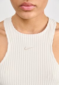Balta juostelėta liemenėlė su aukštu kaklu ir plačiomis petnešomis, kurių priekyje yra mažas šviesiai pilkas išsiuvinėtas Nike logotipas.