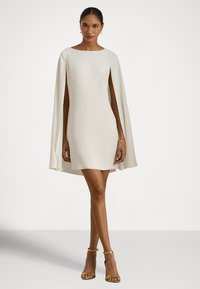 Baltos spalvos trumpa suknelė su valtine iškirpte ir apsiausto formos rankovėmis. Lygus audinys, minimalistinis dizainas ir praplatėjanti siluetas. Aukso spalvos aukštakulniai sandalai.