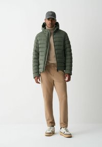 Olíva zöld puffernadrág, bézs bordás pulóver, barna nadrág és fehér sportcipő szürke részletekkel. A modellen sima háttér előtt áll.