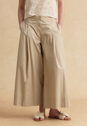 Person trägt beige weite, gekürzte Hose, cremefarbenes Spitzentop und cremefarbene offene Sandalen mit Blumenverzierung, Hände in den Taschen.
