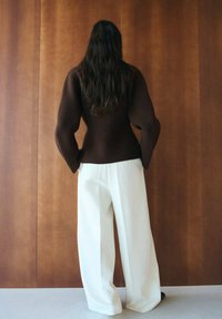 Person mit langen dunklen Haaren steht mit dem Gesicht zur Holzwand, trägt ein dunkelbraunes strukturiertes Oberteil und lockere weiße Hosen mit weitem Bein.