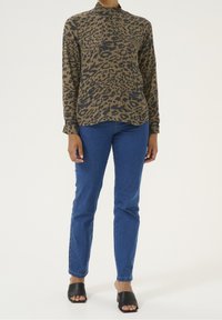 Langærmet bluse med leopardprint i tan og mørkebrun, med høj krave og tekstureret stof, kombineret med blå tætsiddende jeans og sorte åbne tå-mules.