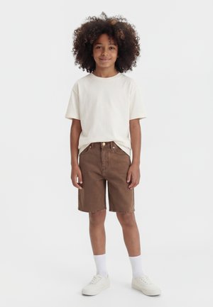 Barn med krøllet hår iført en hvid t-shirt, brune knælange shorts, hvide sokker og hvide sneakers, der står på en ensfarvet baggrund.