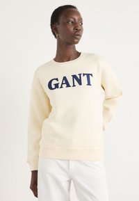 Krémová mikina s vroubkovaným dekoltem a manžetami, na přední straně se nachází velké navrhované logo "GANT" v tmavě modré barvě. Měkká textura.