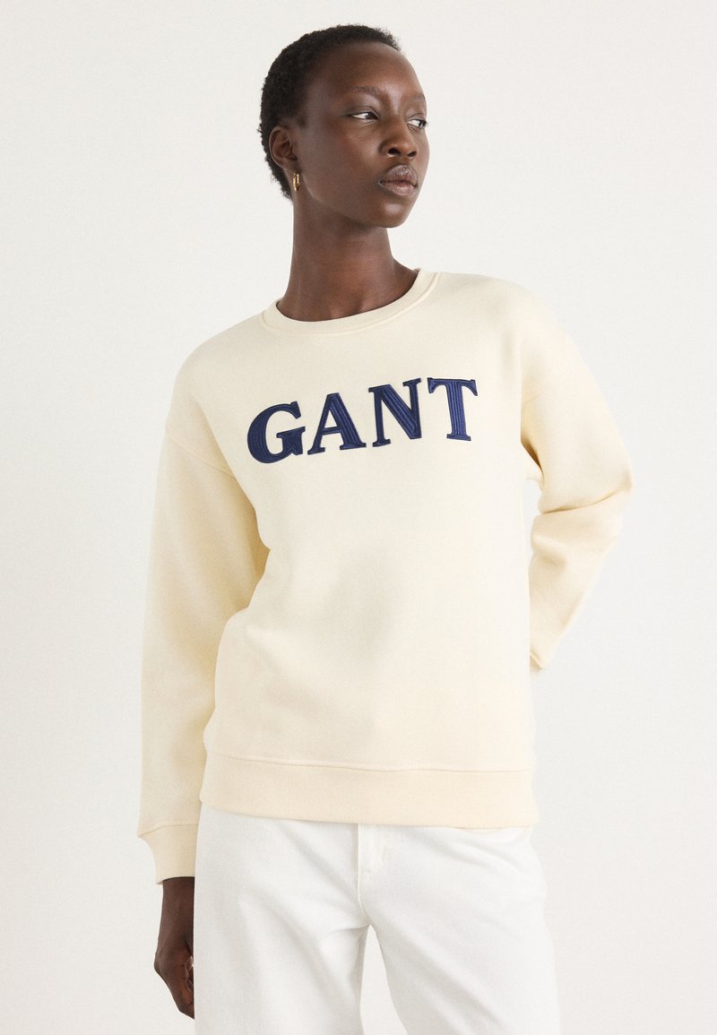 Crème-kleurige sweatshirt met een geribbelde halslijn en boorden, voorzien van een groot marineblauw "GANT"-logo op de voorkant. Zachte textuur.