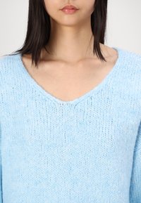 Femme portant un pull en maille duveteuse bleu clair avec un col en V, dévoilant son cou et le haut de sa poitrine, aux cheveux longs et droits foncés.
