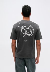 Camiseta de algodón gris con un gran diseño gráfico blanco en la parte posterior que presenta formas abstractas y texto. Mangas cortas, ajuste relajado.