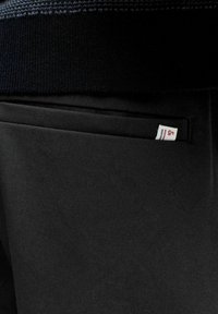 Gros plan sur la poche d'un pantalon noir avec une petite étiquette blanche comportant des rayures rouges et bleues cousue dans la couture de la poche sous l'ourlet d'un pull en maille foncée.