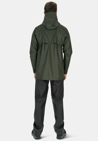 Groene waterdichte jas met capuchon, met lange mouwen en een verstelbare onderrand. Gecombineerd met zwarte waterdichte broek en donkere schoenen.