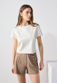 Bílý top s krátkým rukávem a detailem švu, spárovaný s šedobéžovými šortkami, které mají zlaté knoflíkové akcenty na stranách. Hladká textilie.