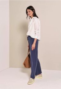 Vit skjorta med knappar, marinblå vida byxor, olivgröna sneakers och brun handväska. Modellen poserar på sidan mot en neutral bakgrund.