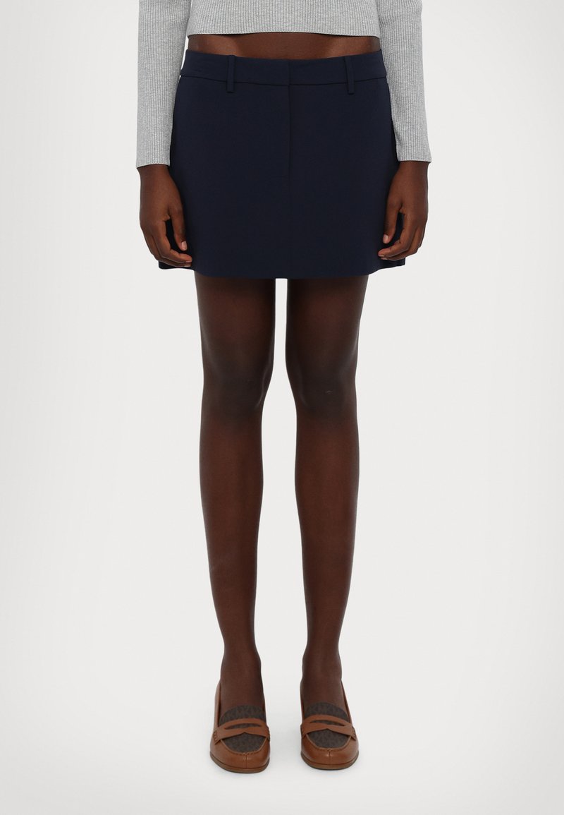 Fustă mini bleumarin cu un design tailoring, față netedă și buzunare laterale subtile. Purtați cu pantofi maro cu detalii texturate.
