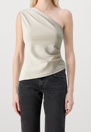 Kvinde iført en lysebeige, enkelt-ærmet, ærmeløs top og højtaljede sorte jeans, stående foran en ensfarvet baggrund.