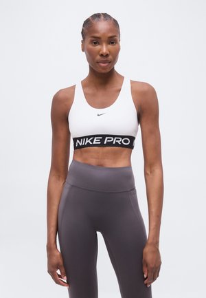 Mujer atlética con el cabello trenzado lleva un sujetador deportivo blanco de Nike y leggings grises de talle alto, de pie frente a un fondo blanco liso.