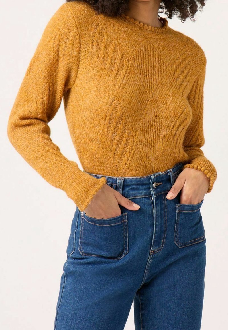 Musztardowożółty sweter dziany o fakturze w diamentowy wzór, z okrągłym dekoltem i ściągaczami przy rękawach, zestawiony z niebieskimi jeansami z wysokim stanem.