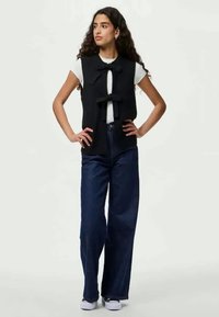 Colete preto sem mangas com detalhe de laço, usado sobre uma camisola branca, combinado com calças de ganga largas de denim escuro e sapatilhas brancas.