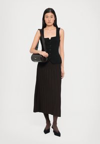Black sleeveless vest with buttons paired with a pleated dark brown midi skirt. Model holds a round black handbag with a shoulder strap.