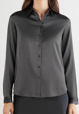 Woman wearing a long-sleeve, buttoned dark gray silk blouse with a collar, paired with a dark skirt against a plain background.