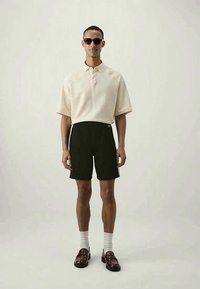 Camisa polo creme com mangas curtas, calções pretos, meias brancas e mocassins castanhos; indivíduo a usar óculos de sol, de pé contra um fundo neutro.
