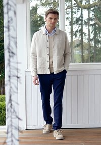 Giacca beige con bottoni sopra un maglione crema, pantaloni blu scuro e sneakers beige chiaro. In piedi contro un muro bianco con vegetazione all'esterno.