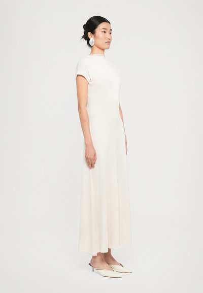 Vrouw in een off-white maxi-jurk met korte mouwen en een hoge hals, gecombineerd met off-white muiltjes met een spitse neus en grote oorbellen.