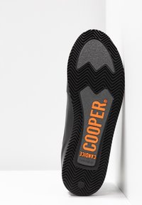 Černá gumová podrážka boty má vzor špičatých zubů ve tvaru cik cak, s nápisem "COOPER" v tučném oranžovém písmu na šedém pozadí.