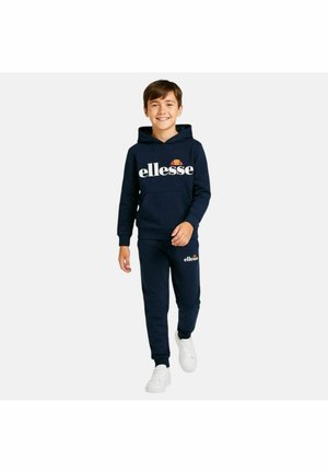 Garçon marchant vers l'avant, portant un sweat à capuche ellesse bleu marine et un pantalon de survêtement, avec des baskets blanches, souriant devant un fond clair uni.