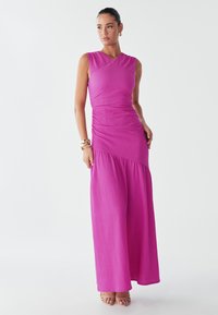 Model poses in a vibrant pink, sleeveless dress with ruched detailing and a flowing skirt. She wears gold accessories and stands against a light backdrop.