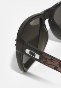 Sorte solbriller med mørke linser, teksturerte armer med et rødt grafisk design og sølv detaljer. Kompakt, sporty fasong.