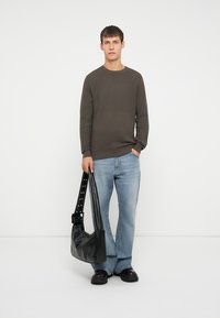 Suéter de patrón marrón oscuro, jeans azul claro y un bolso negro con correa con tachuelas, usado por un modelo que se encuentra frente a un fondo liso.