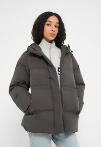 Mujer vestida con una chaqueta de plumón gris oscuro, con las manos en los bolsillos, sudadera con cremallera gris claro y jeans azul claro, de pie contra un fondo blanco.