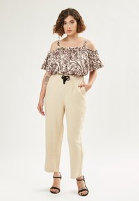Off-shoulder top in een bruine en crèmekleurige abstracte print, met ruches; crèmekleurige broek met elastische taille en zwarte koord; zwarte sandalen.