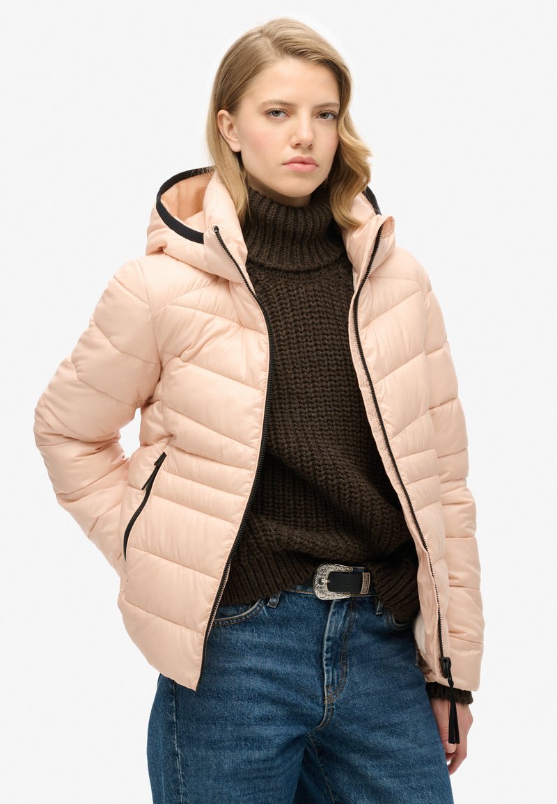 Giacca puffa rosa chiaro con cappuccio, con dettagli neri e chiusura a zip. Indossata sopra un maglione lavorato a maglia marrone scuro e jeans blue.