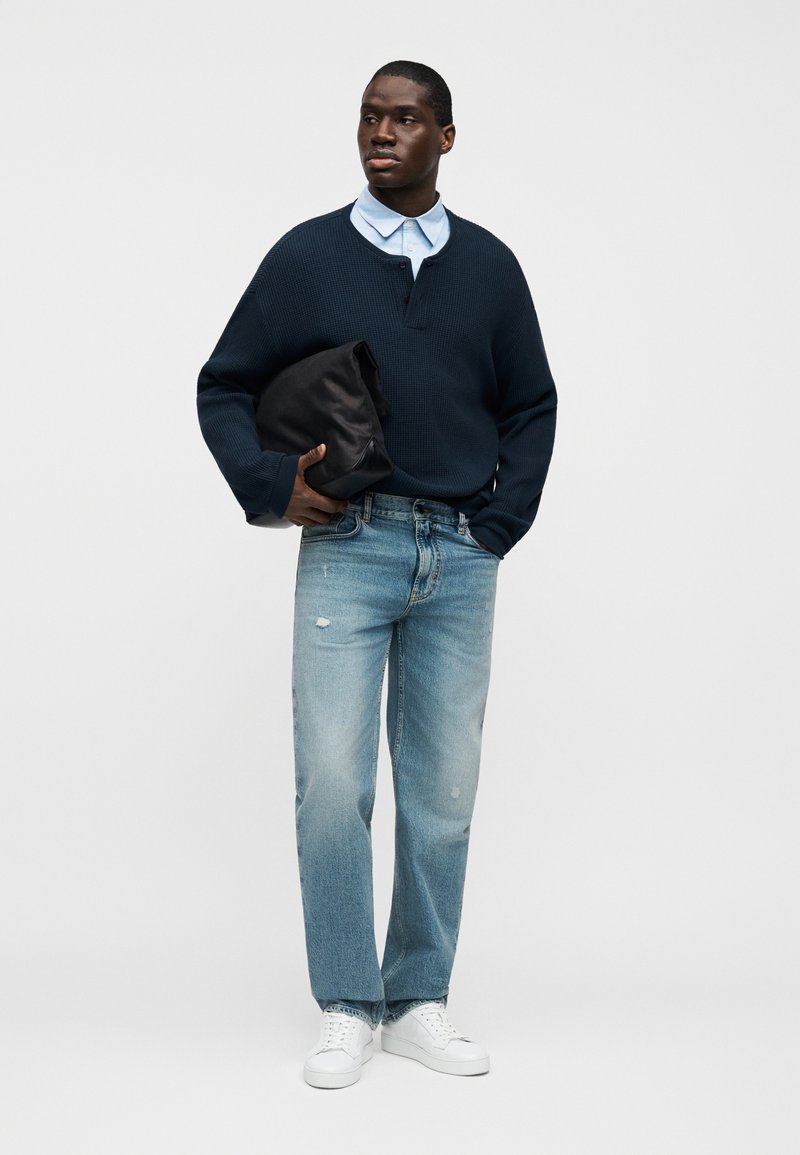 Pull-over en maille texturée bleu marine avec un col boutonné, chemise bleu clair en dessous, jean bleu clair déchiré, et baskets blanches. Tenant un sac noir.