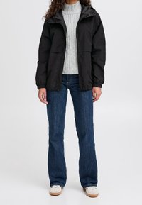 Svart jacka med huva, med dragkedjestängning och elastiska ärmslut, bärs över en ljusgrå tröja och blå jeans. Vita sneakers med beige sålar.