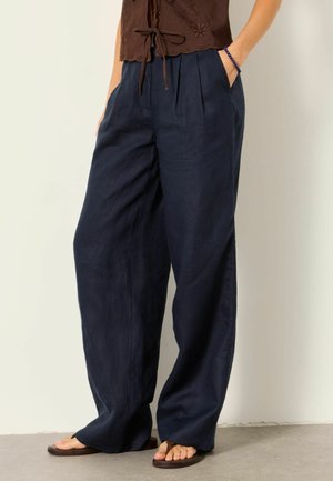 Nainen, jolla on päällään väljät tummansiniset pellavahousut laskoksilla, ruskea hihaton toppi, sininen helmirannekoru ja ruskeat varvasremmillä olevat sandaalit.